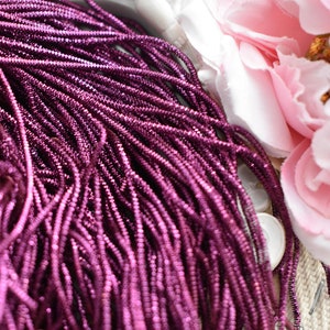 May include: A close-up of a bundle of metallic purple seed beads. The beads are small and round, and they have a shiny finish.