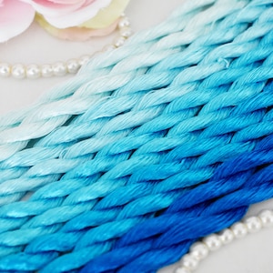 May include: A collection of blue and white embroidery floss spools, arranged in a gradient from white to dark blue. The floss is braided and has a soft, silky texture.