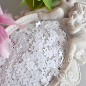 Op de afbeelding: Een decoratief dienblad gevuld met witte, bloemvormige confetti, vastgehouden door een wit cherubijnbeeld. Kunstmatige roze en groene bloemen zijn op de achtergrond. Deze confetti is geschikt voor knutselen, scrapbooking of feestversieringen.