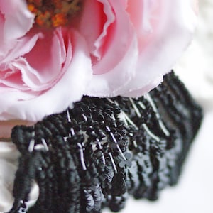 Może przedstawiać: Różowa róża z tkaniny z czarnym koralikowym wykończeniem. Róża jest wykonana z miękkiej tkaniny i ma delikatną, falbaniastą fakturę.