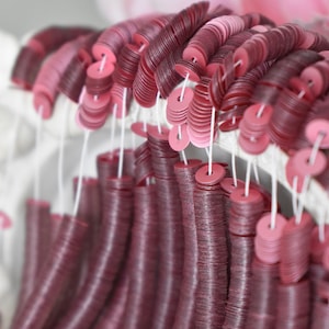 Pode incluir: Close-up de uma coleção de lantejoulas bordô e rosa enfiadas. As lantejoulas são circulares com um furo no centro, criando um efeito texturizado e em camadas. A disposição sugere um item artesanal ou decorativo.
