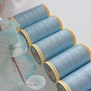 May include: A row of six spools of light blue thread, each with a yellow label. The spools are arranged in a diagonal line, with the top spool slightly overlapping the one below it.