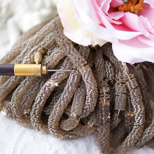 Może przedstawiać: Zbliżenie dekoracyjnego przedmiotu z ciemnym drewnianym uchwytem i złotymi metalowymi akcentami. Obiekt ma skupisko brązowych, teksturowanych pasm. Różowy i biały kwiat jest w tle.