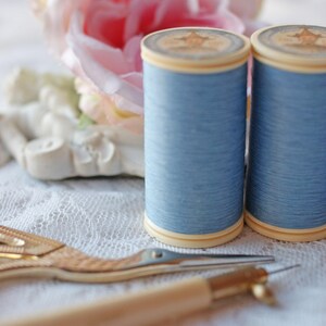 Op de afbeelding: Twee spoelen lichtblauw garen liggen naast een goudkleurige borduurschaar en een naai-instrument met houten handvat. Een roze roos en een wit decoratief element staan op de achtergrond. De items staan op een wit kanten oppervlak.