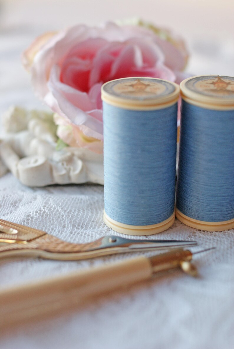 Op de afbeelding: Twee spoelen lichtblauw garen liggen naast een goudkleurige borduurschaar en een naai-instrument met houten handvat. Een roze roos en een wit decoratief element staan op de achtergrond. De items staan op een wit kanten oppervlak.
