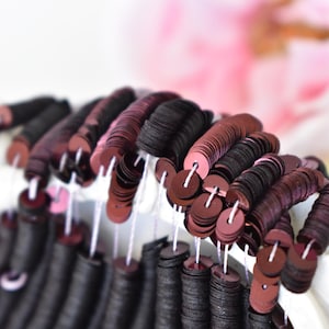 Puede incluir: Primer plano de una gran cantidad de lentejuelas redondas negras y burdeos, enhebradas en un hilo blanco. Las lentejuelas están dispuestas en filas, creando un efecto de cascada.