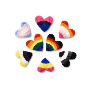 Op de afbeelding: Zeven gehaakte harten in verschillende kleuren en patronen, waaronder regenboog-, transgender- en biseksuele regenboogvlaggen, evenals zwart-wit en roze en geel.