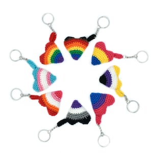 Op de afbeelding: Een set van negen gebreide hartvormige sleutelhangers, elk met een andere pride-vlag. De sleutelhangers zijn in een cirkel op een witte achtergrond gerangschikt.