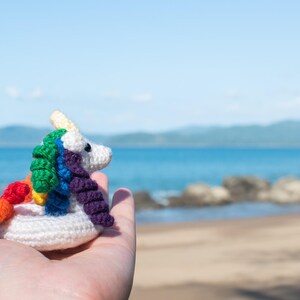 Op de afbeelding: Een gehaakt regenboog unicorn zwembandje, vastgehouden in een hand tegen een strandachtergrond.