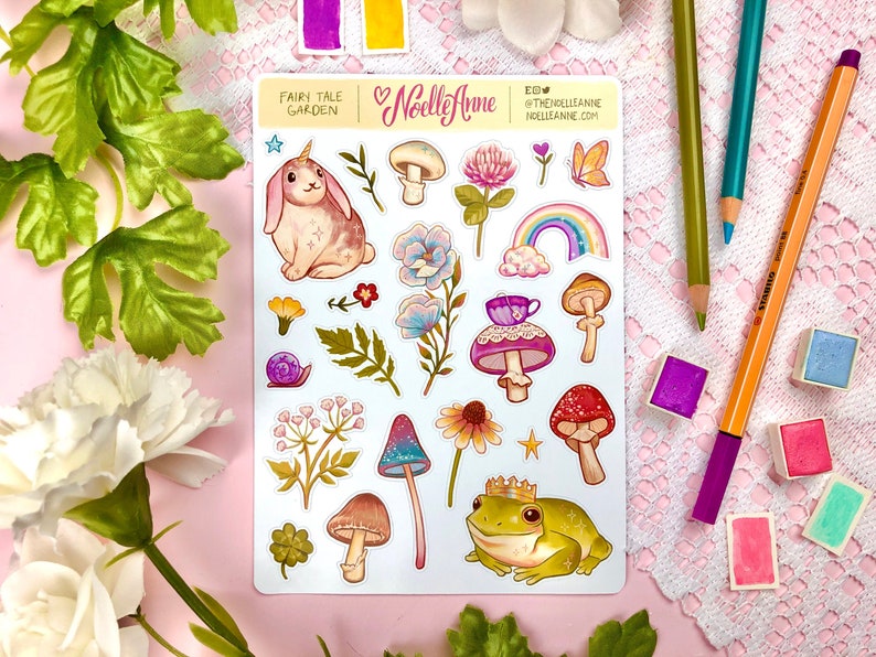 May include: A sheet of stickers featuring a whimsical fairy tale garden theme. The stickers include a unicorn bunny, mushrooms, flowers, a frog wearing a crown, a teacup, and a rainbow. The stickers are printed on white paper with a glossy finish.