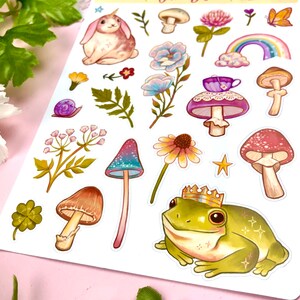 May include: A sheet of colorful stickers featuring whimsical illustrations of a unicorn rabbit, mushrooms, flowers, a frog wearing a crown, and a snail. The stickers are perfect for adding a touch of whimsy to journals, planners, and other craft projects.