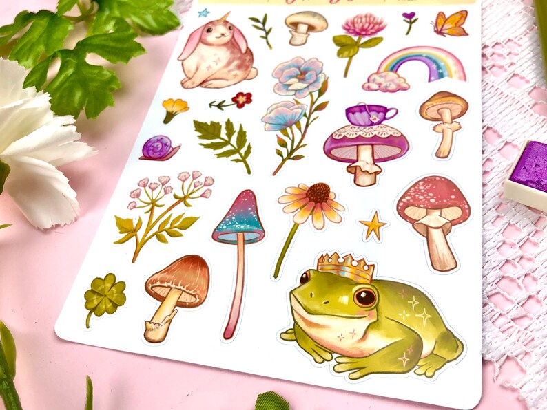 May include: A sheet of colorful stickers featuring whimsical illustrations of a unicorn rabbit, mushrooms, flowers, a frog wearing a crown, and a snail. The stickers are perfect for adding a touch of whimsy to journals, planners, and other craft projects.