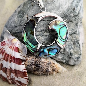May include: A silver pendant necklace featuring a circular abalone shell design. The shell displays a swirling pattern of blue, green, and iridescent colours. The pendant is resting on a grey rock and a seashell.