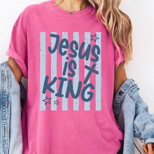 May include: A bright pink t-shirt featuring the words "Jesus is King" in blue script, set against a backdrop of vertical blue stripes. The shirt has rolled-up sleeves and a relaxed fit. The word "Crunchberry" is in the top left corner.