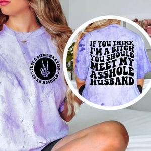 May include: A light purple tie-dye t-shirt with black text and graphics. The front features a circle with the text "Just a Bitch Married to an Asshole" and a hand making a peace sign with a skeleton hand. The back says "If you think I'm a bitch you should meet my asshole husband."