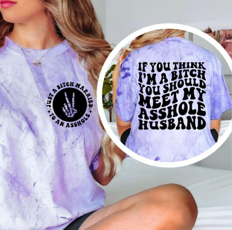 May include: A light purple tie-dye t-shirt with black text and graphics. The front features a circle with the text "Just a Bitch Married to an Asshole" and a hand making a peace sign with a skeleton hand. The back says "If you think I'm a bitch you should meet my asshole husband."