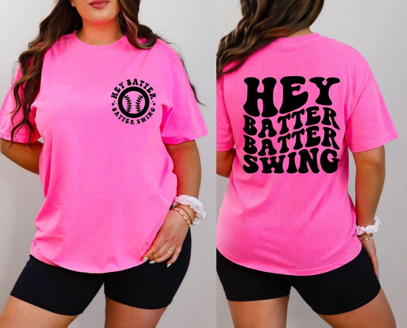 K&ouml;nnte beinhalten: Ein leuchtend pinkfarbenes T-Shirt mit dem schwarzen Schriftzug "HEY BATTER BATTER SWING" auf dem R&uuml;cken. Auf der Vorderseite befindet sich eine Baseball-Grafik mit dem gleichen Text in einem Kreis. Das Shirt wird mit schwarzen Shorts getragen.