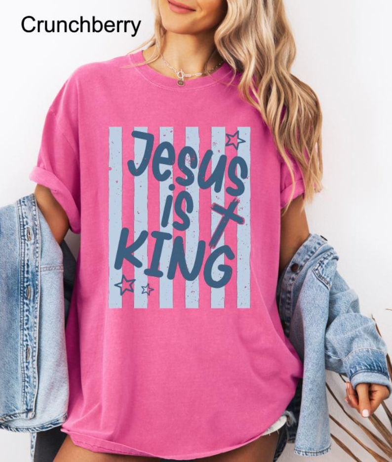 May include: A bright pink t-shirt featuring the words "Jesus is King" in blue script, set against a backdrop of vertical blue stripes. The shirt has rolled-up sleeves and a relaxed fit. The word "Crunchberry" is in the top left corner.