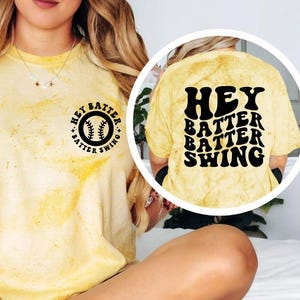 K&ouml;nnte beinhalten: Gelbes T-Shirt mit Batikmuster und dem Text "Hey Batter Batter Shing" in Schwarz auf dem R&uuml;cken. Auf der Vorderseite befindet sich eine Baseball-Grafik mit dem Text "Hey Batter Batter Swing" in einem kreisf&ouml;rmigen Design.