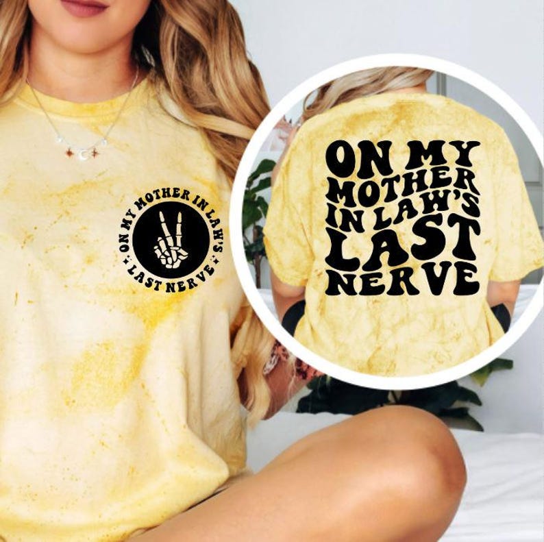 May include: Yellow tie-dye t-shirt with black text and graphics. The front features a circular design with a skeleton hand making a peace sign. The back of the shirt has the phrase "ON MY MOTHER IN LAW'S LAST NERVE" in large, bold letters.