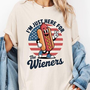 May include: Ivory-colored t-shirt with a cartoon hot dog character giving a peace sign. The design includes the text "I'M JUST HERE FOR The Wieners" with an American flag background. The hot dog is wearing blue shoes.