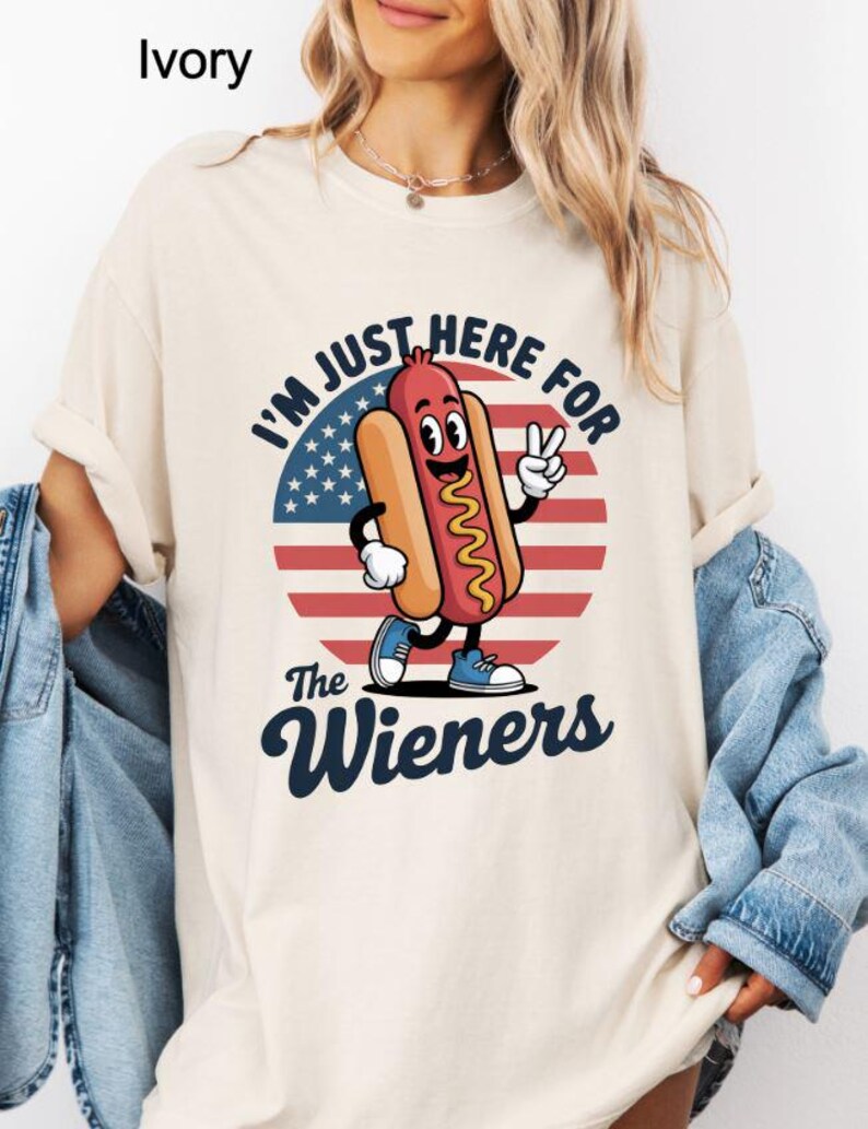 May include: Ivory-colored t-shirt with a cartoon hot dog character giving a peace sign. The design includes the text "I'M JUST HERE FOR The Wieners" with an American flag background. The hot dog is wearing blue shoes.
