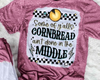 May include: A maroon t-shirt with a bleached, speckled design. The shirt features a graphic with the text "Some of y'alls CORNBREAD ain't done in the MIDDLE" with a cornbread illustration and checkered accents.