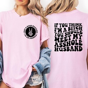 May include: A light pink t-shirt with the text "JUST A BITCH MARRIED TO AN ASSHOLE" on the front and "IF YOU THINK I'M A BITCH YOU SHOULD MEET MY ASSHOLE HUSBAND" on the back. The front graphic is a circle with a peace sign made of a skeleton hand.