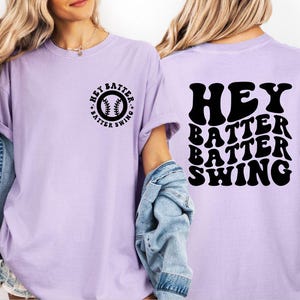 K&ouml;nnte beinhalten: Helllila T-Shirt mit dem schwarzen Schriftzug "HEY BATTER SWING". Auf der Vorderseite des Shirts befindet sich ein kleines kreisf&ouml;rmiges Design mit einem Baseball und demselben Text. Das Shirt hat kurze &Auml;rmel und eine lockere Passform.