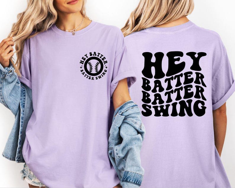 K&ouml;nnte beinhalten: Helllila T-Shirt mit dem schwarzen Schriftzug "HEY BATTER SWING". Auf der Vorderseite des Shirts befindet sich ein kleines kreisf&ouml;rmiges Design mit einem Baseball und demselben Text. Das Shirt hat kurze &Auml;rmel und eine lockere Passform.