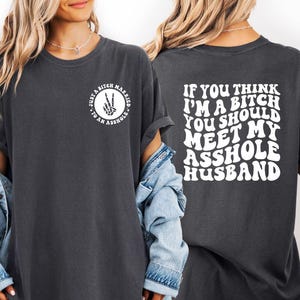 May include: Dark gray t-shirt with white text on the front and back. The front reads "Just a Bitch Married to an Asshole" in a circle with a peace sign. The back says "If you think I'm a bitch you should meet my asshole husband."