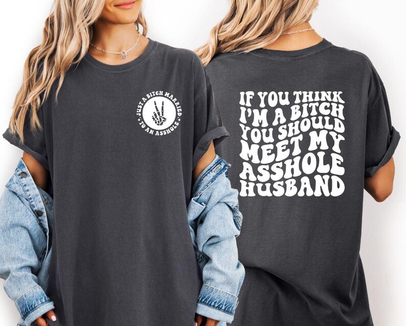 May include: Dark gray t-shirt with white text on the front and back. The front reads "Just a Bitch Married to an Asshole" in a circle with a peace sign. The back says "If you think I'm a bitch you should meet my asshole husband."