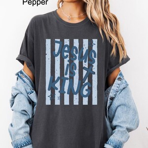 May include: Dark grey t-shirt featuring the text "Jesus is King" in blue. The text is set against a backdrop of vertical light blue stripes. The shirt has short sleeves and a relaxed fit.