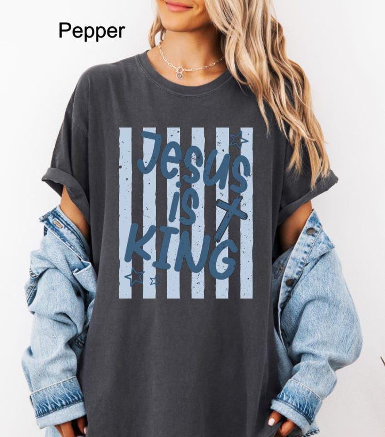 May include: Dark grey t-shirt featuring the text "Jesus is King" in blue. The text is set against a backdrop of vertical light blue stripes. The shirt has short sleeves and a relaxed fit.