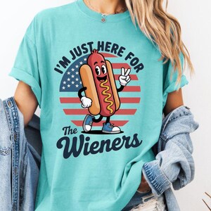 May include: A turquoise t-shirt with the text "I'M JUST HERE FOR The Wieners" and a cartoon hot dog character. The hot dog is smiling and giving a peace sign in front of a red, white, and blue graphic.