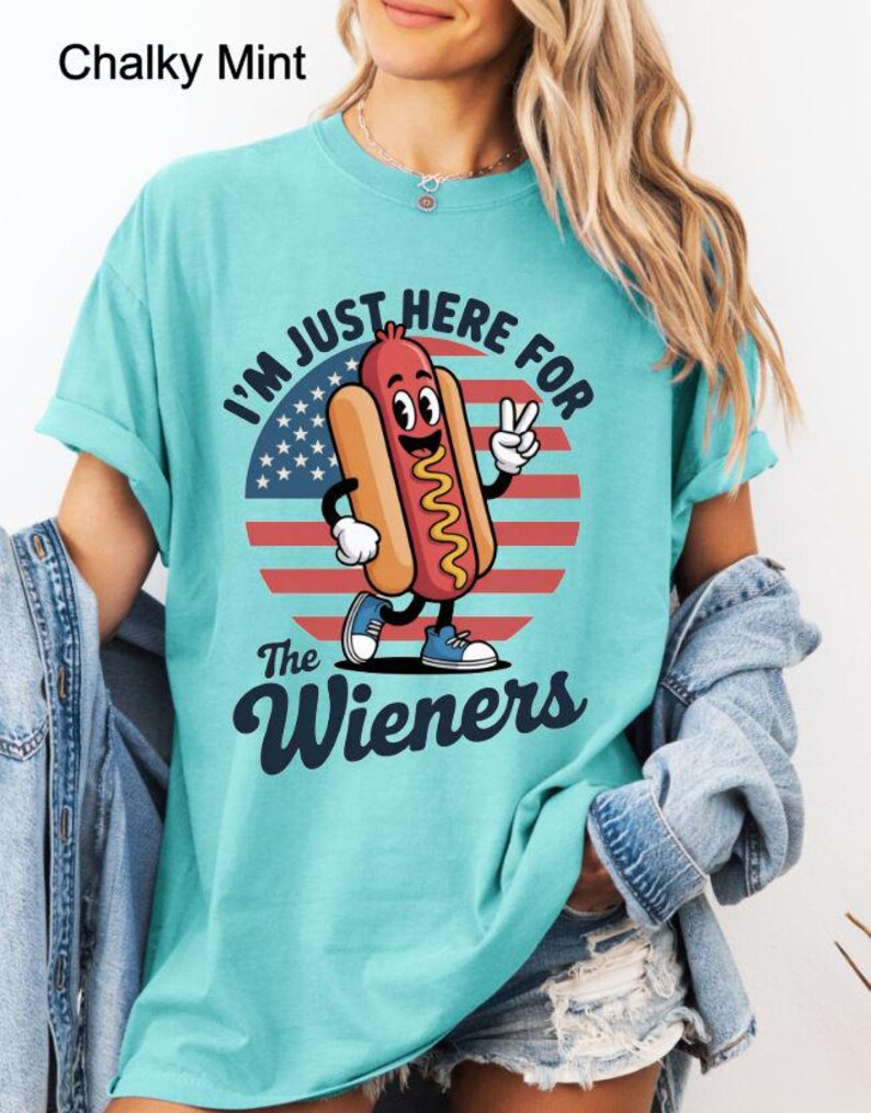 May include: A turquoise t-shirt with the text "I'M JUST HERE FOR The Wieners" and a cartoon hot dog character. The hot dog is smiling and giving a peace sign in front of a red, white, and blue graphic.