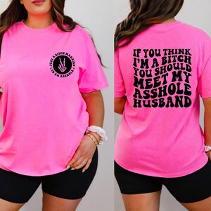 May include: Bright pink t-shirt with black text on the front and back. The front reads "Just a bitch married to an asshole" with a peace sign made of bones. The back says "If you think I'm a bitch you should meet my asshole husband."