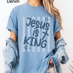 May include: A light blue t-shirt with the text "Jesus is King" in a blue and white design. The shirt has a washed denim appearance and is worn with a denim jacket.