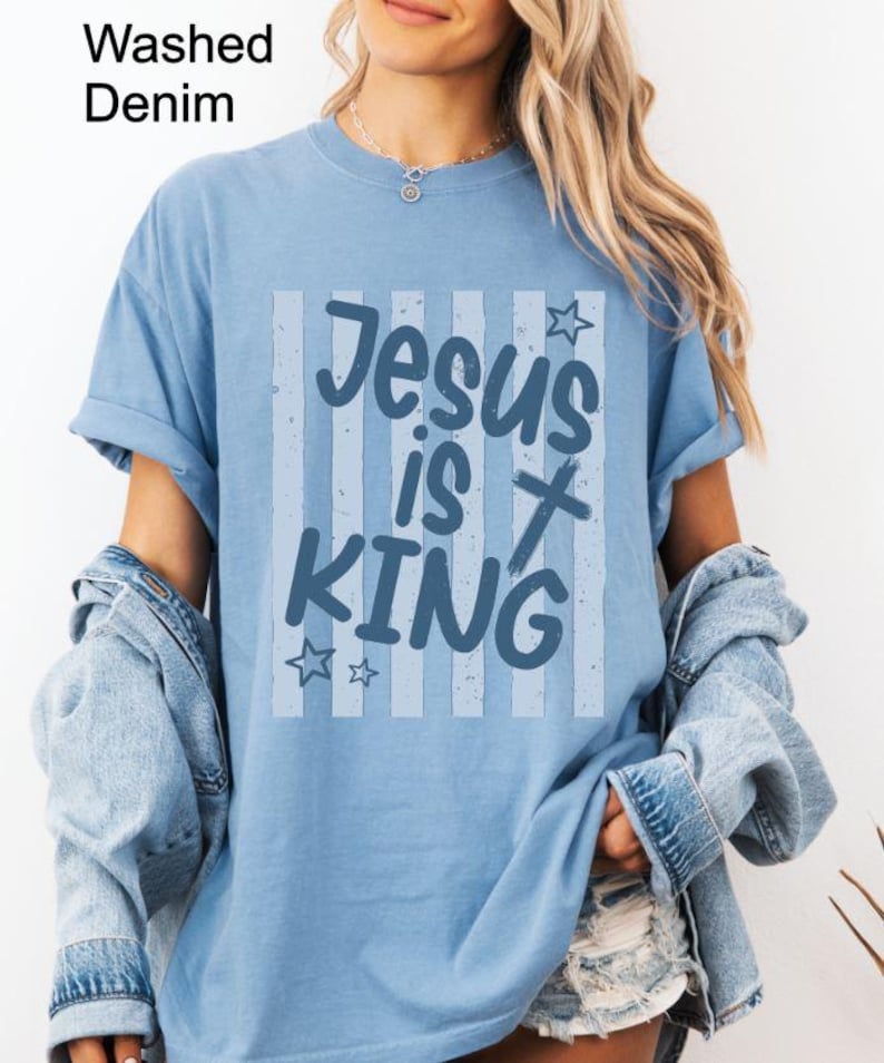 May include: A light blue t-shirt with the text "Jesus is King" in a blue and white design. The shirt has a washed denim appearance and is worn with a denim jacket.