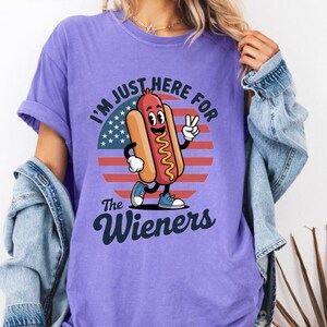 May include: Violet-colored t-shirt with a graphic of a cartoon hot dog giving a peace sign in front of a red, white, and blue background. The text reads "I'M JUST HERE FOR THE Wieners."