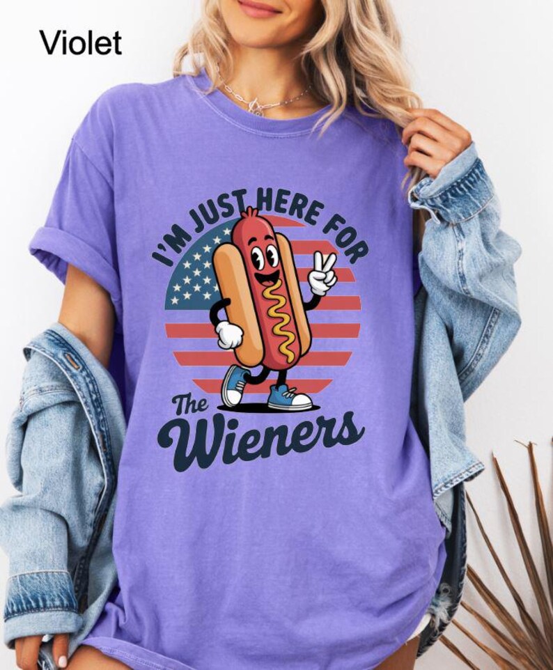 May include: Violet-colored t-shirt with a graphic of a cartoon hot dog giving a peace sign in front of a red, white, and blue background. The text reads "I'M JUST HERE FOR THE Wieners."