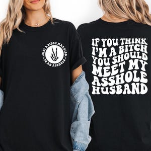 May include: Black t-shirt with white text on the front and back. The front features a circle with a peace sign made of bones and the words "Just a Bitch Married to an Asshole." The back says, "If you think I'm a bitch you should meet my asshole husband."