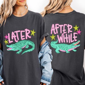 May include: Dark gray t-shirt with a teal alligator graphic and the word "LATER" in pink letters with yellow and pink stars. The back of the shirt says "AFTER WHILE" with the same alligator graphic.