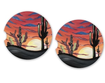 Puede incluir: Dos posavasos redondos para coche con un diseño de atardecer en el desierto. El diseño incluye cactus, un sol poniente y un cielo vibrante con tonos rojos, naranjas y azules. Los posavasos tienen un borde negro y un recorte.