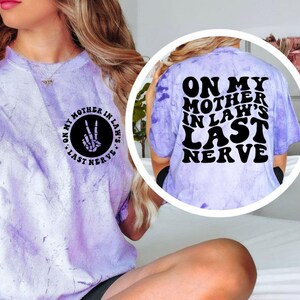 May include: A light purple tie-dye t-shirt with the text "ON MY MOTHER IN LAW'S LAST NERVE" in black. The front of the shirt has a peace sign made of bones. The back of the shirt has the same text in a larger font.