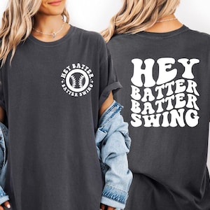 K&ouml;nnte beinhalten: Dunkelgraues T-Shirt mit dem wei&szlig;en Schriftzug "Hey Batter Batter Swing". Auf der Vorderseite befindet sich ein kreisf&ouml;rmiges Design mit einem Baseball und demselben Text. Das T-Shirt hat kurze &Auml;rmel und eine lockere Passform.