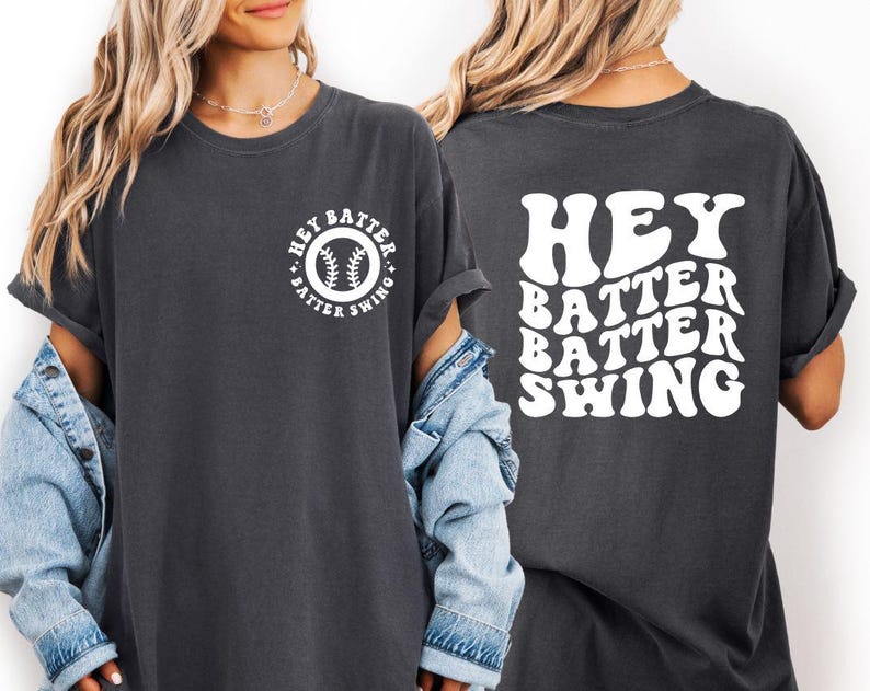 K&ouml;nnte beinhalten: Dunkelgraues T-Shirt mit dem wei&szlig;en Schriftzug "Hey Batter Batter Swing". Auf der Vorderseite befindet sich ein kreisf&ouml;rmiges Design mit einem Baseball und demselben Text. Das T-Shirt hat kurze &Auml;rmel und eine lockere Passform.