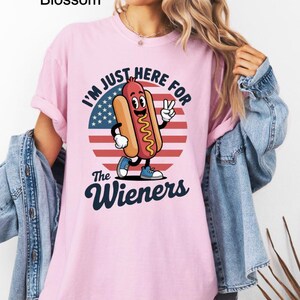 May include: A light pink t-shirt with the text "I'M JUST HERE FOR The Wieners" and a cartoon hot dog character. The hot dog is wearing blue shoes and giving a peace sign. The background is a circle with the American flag.