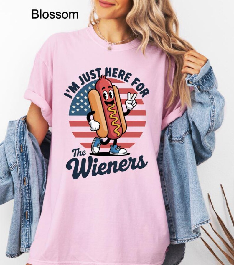 May include: A light pink t-shirt with the text "I'M JUST HERE FOR The Wieners" and a cartoon hot dog character. The hot dog is wearing blue shoes and giving a peace sign. The background is a circle with the American flag.
