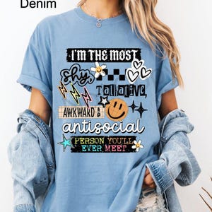 K&ouml;nnte beinhalten: Hellblaues T-Shirt mit dem Text "I'M THE MOST shy, talkative, awkward, antisocial PERSON YOU'LL EVER MEET" in verschiedenen Schriftarten und Grafiken. Das T-Shirt hat einen Washed-Denim-Look.